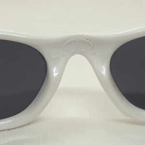 Puede incluir: Primer plano de gafas de sol blancas con lentes oscuras. Las gafas de sol tienen una montura redondeada y un peque&ntilde;o emblema en el puente. Las lentes son de forma ovalada y el dise&ntilde;o general es elegante y moderno.