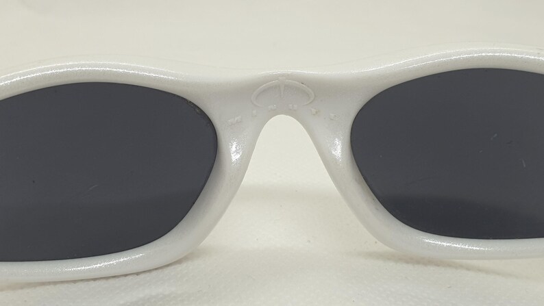 Puede incluir: Primer plano de gafas de sol blancas con lentes oscuras. Las gafas de sol tienen una montura redondeada y un peque&ntilde;o emblema en el puente. Las lentes son de forma ovalada y el dise&ntilde;o general es elegante y moderno.