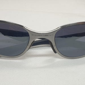 K&ouml;nnte beinhalten: Silberfarbene Sonnenbrille mit dunkelgrauen Gl&auml;sern. Die Sonnenbrille hat ein geschwungenes Design, und der Markenname "Oakley" ist auf dem Steg sichtbar. Die B&uuml;gel sind schwarz und haben ein abgerundetes Ende.