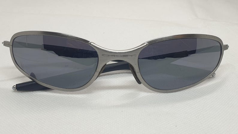 K&ouml;nnte beinhalten: Silberfarbene Sonnenbrille mit dunkelgrauen Gl&auml;sern. Die Sonnenbrille hat ein geschwungenes Design, und der Markenname "Oakley" ist auf dem Steg sichtbar. Die B&uuml;gel sind schwarz und haben ein abgerundetes Ende.