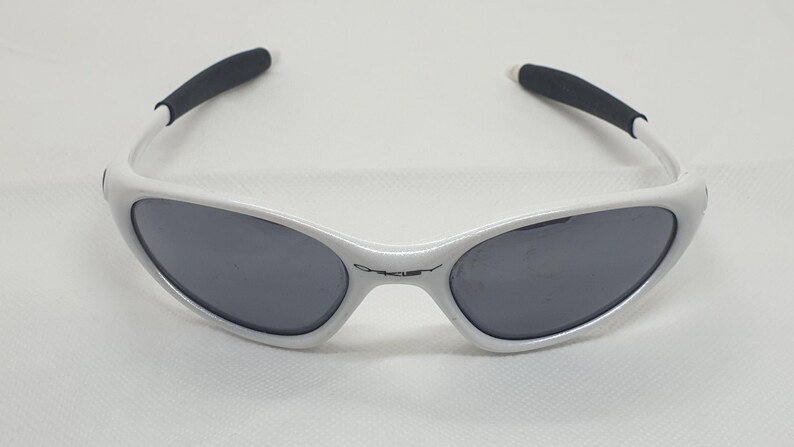 Puede incluir: Gafas de sol blancas con lentes grises oscuras y puntas de patillas negras. Las gafas de sol tienen un dise&ntilde;o elegante y moderno, con el nombre de la marca sutilmente mostrado en la montura.