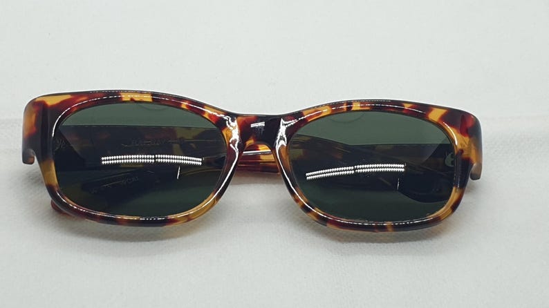 Puede incluir: Gafas de sol con montura de carey y lentes verde oscuro. Las monturas tienen una forma redondeada cl&aacute;sica con un patr&oacute;n moteado marr&oacute;n y &aacute;mbar. Las lentes son de color verde oscuro, contrastando con la montura.