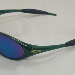 May include: Green sunglasses with a glossy finish and blue-purple mirrored lenses. The frames have a curved design with black rubberized ear pieces. The Oakley logo is visible on the temples.
