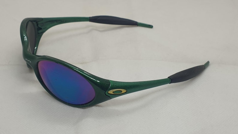 May include: Green sunglasses with a glossy finish and blue-purple mirrored lenses. The frames have a curved design with black rubberized ear pieces. The Oakley logo is visible on the temples.