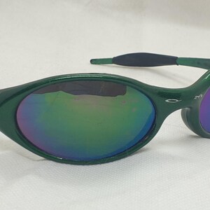 May include: Green sunglasses with a curved frame and iridescent lenses. The lenses reflect a spectrum of colors, including blue, green, and purple. The sunglasses have a sleek, modern design, with a small gold emblem on the frame.