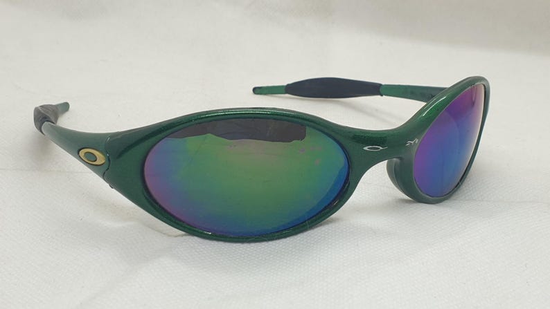 May include: Green sunglasses with a curved frame and iridescent lenses. The lenses reflect a spectrum of colors, including blue, green, and purple. The sunglasses have a sleek, modern design, with a small gold emblem on the frame.