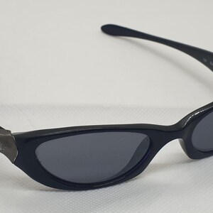 May include: Black sunglasses with a curved frame and dark grey lenses. The sunglasses have a small, metallic emblem on the side of the frame. The arms curve back towards the ears.