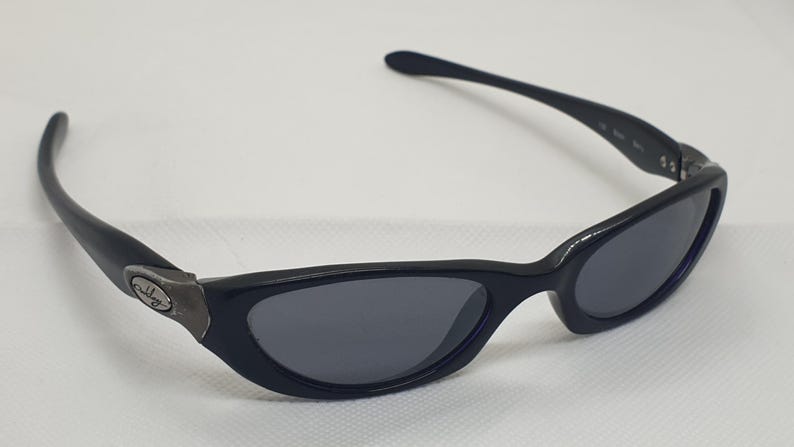 May include: Black sunglasses with a curved frame and dark grey lenses. The sunglasses have a small, metallic emblem on the side of the frame. The arms curve back towards the ears.