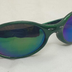 May include: Green-framed sunglasses with iridescent lenses. The sunglasses have a curved design and a glossy finish. The lenses reflect a spectrum of colors, including blue, green, and purple. The frame has a small logo on the temple.