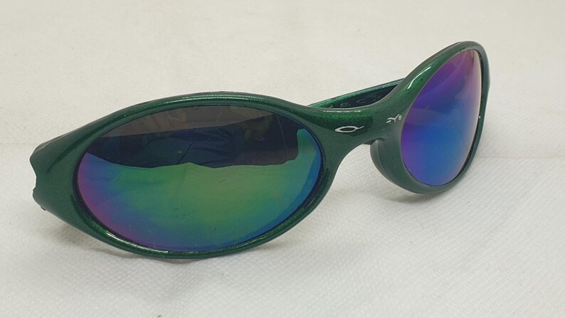 May include: Green-framed sunglasses with iridescent lenses. The sunglasses have a curved design and a glossy finish. The lenses reflect a spectrum of colors, including blue, green, and purple. The frame has a small logo on the temple.