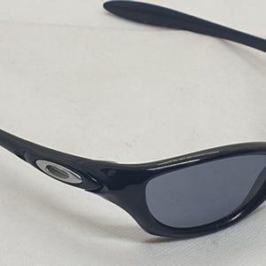 Puede incluir: Gafas de sol negras con un dise&ntilde;o elegante y curvado. Las gafas tienen lentes de color gris oscuro y una montura negra con un logotipo plateado en las patillas. El estilo es deportivo y moderno.