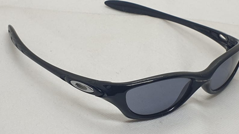 Puede incluir: Gafas de sol negras con un dise&ntilde;o elegante y curvado. Las gafas tienen lentes de color gris oscuro y una montura negra con un logotipo plateado en las patillas. El estilo es deportivo y moderno.