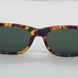Puede incluir: Gafas de sol con montura con estampado de carey y lentes verde oscuro. Las gafas de sol tienen una forma rectangular cl&aacute;sica con una l&iacute;nea de cejas ligeramente curvada. Las patillas son de color marr&oacute;n oscuro.