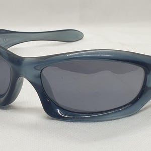 May include: A pair of blue-grey sunglasses with dark grey lenses. The sunglasses have a curved frame and a small logo on the temple. The lenses are oval-shaped, and the frame is made of a glossy plastic material.