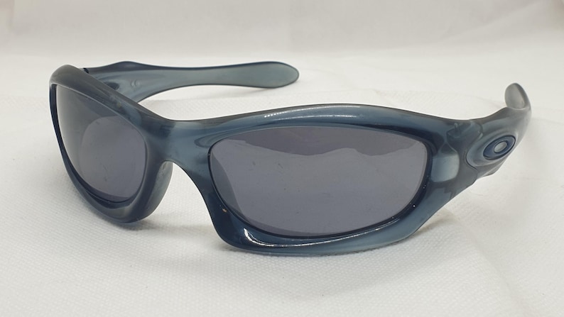 May include: A pair of blue-grey sunglasses with dark grey lenses. The sunglasses have a curved frame and a small logo on the temple. The lenses are oval-shaped, and the frame is made of a glossy plastic material.