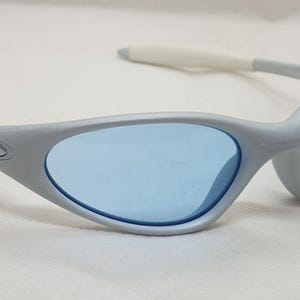 K&ouml;nnte beinhalten: Graue Sonnenbrille mit blauen Gl&auml;sern. Die Sonnenbrille hat ein geschwungenes, umlaufendes Design mit wei&szlig;en Akzenten an den B&uuml;geln. Das Oakley-Logo ist auf der Seite des Rahmens sichtbar. Die Gl&auml;ser sind hellblau.