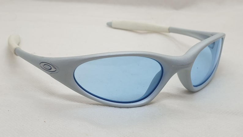 K&ouml;nnte beinhalten: Graue Sonnenbrille mit blauen Gl&auml;sern. Die Sonnenbrille hat ein geschwungenes, umlaufendes Design mit wei&szlig;en Akzenten an den B&uuml;geln. Das Oakley-Logo ist auf der Seite des Rahmens sichtbar. Die Gl&auml;ser sind hellblau.