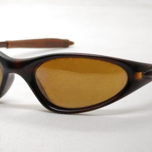 Puede incluir: Gafas de sol ovaladas marrones con lentes &aacute;mbar y montura marr&oacute;n oscuro. Las patillas son de un marr&oacute;n m&aacute;s claro. Las gafas de sol est&aacute;n sobre un fondo blanco.
