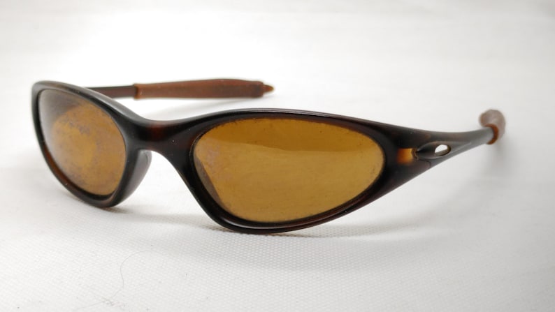 Puede incluir: Gafas de sol ovaladas marrones con lentes &aacute;mbar y montura marr&oacute;n oscuro. Las patillas son de un marr&oacute;n m&aacute;s claro. Las gafas de sol est&aacute;n sobre un fondo blanco.