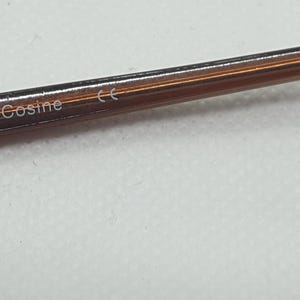 Pode incluir: Close-up de uma arma&ccedil;&atilde;o de &oacute;culos castanha com a palavra "Cosine" impressa na haste. A arma&ccedil;&atilde;o tem um brilho met&aacute;lico e um design elegante e moderno. O fundo &eacute; uma superf&iacute;cie lisa de cor clara.