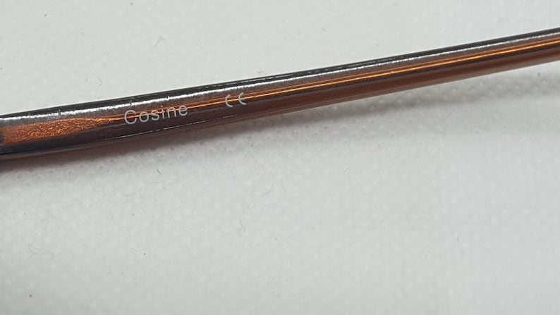 Pode incluir: Close-up de uma arma&ccedil;&atilde;o de &oacute;culos castanha com a palavra "Cosine" impressa na haste. A arma&ccedil;&atilde;o tem um brilho met&aacute;lico e um design elegante e moderno. O fundo &eacute; uma superf&iacute;cie lisa de cor clara.
