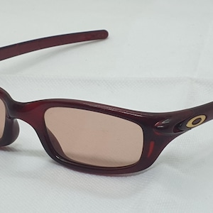 May include: Maroon sunglasses with a rectangular frame and tinted lenses. The temples feature a gold emblem. These glasses are designed for eye protection and fashion.