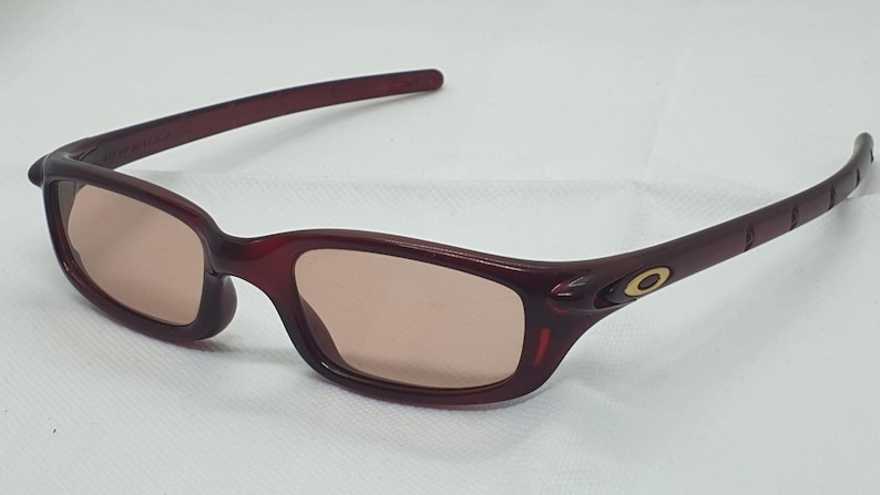 May include: Maroon sunglasses with a rectangular frame and tinted lenses. The temples feature a gold emblem. These glasses are designed for eye protection and fashion.