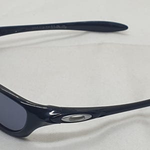 Puede incluir: Gafas de sol negras con un dise&ntilde;o curvo. Las lentes son de color gris oscuro y las monturas son de color negro brillante. El logotipo de Oakley es visible en las patillas. Estas gafas de sol est&aacute;n dise&ntilde;adas para deportes y actividades al aire libre.