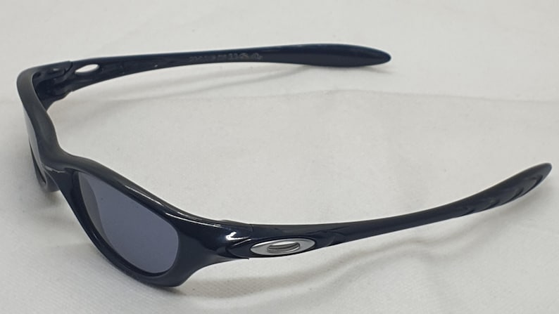 Puede incluir: Gafas de sol negras con un dise&ntilde;o curvo. Las lentes son de color gris oscuro y las monturas son de color negro brillante. El logotipo de Oakley es visible en las patillas. Estas gafas de sol est&aacute;n dise&ntilde;adas para deportes y actividades al aire libre.