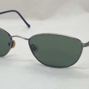 Puede incluir: Gafas de sol con montura plateada y lentes verdes ovaladas. Las monturas tienen un aspecto fino y met&aacute;lico con una sutil curva en las patillas. Las varillas son negras en los extremos. Un accesorio de gafas cl&aacute;sico.