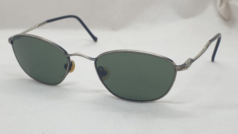 Puede incluir: Gafas de sol con montura plateada y lentes verdes ovaladas. Las monturas tienen un aspecto fino y met&aacute;lico con una sutil curva en las patillas. Las varillas son negras en los extremos. Un accesorio de gafas cl&aacute;sico.