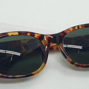 Puede incluir: Gafas de sol carey con lentes verde oscuro. Las monturas presentan un patr&oacute;n cl&aacute;sico de marr&oacute;n, &aacute;mbar y negro. Estas gafas de sol son un accesorio elegante para la protecci&oacute;n de los ojos del sol.