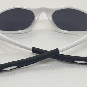Puede incluir: Gafas de sol blancas con lentes grises oscuras. Las gafas tienen una montura y patillas blancas, con agarres de goma negra en los extremos. Las patillas est&aacute;n cruzadas.