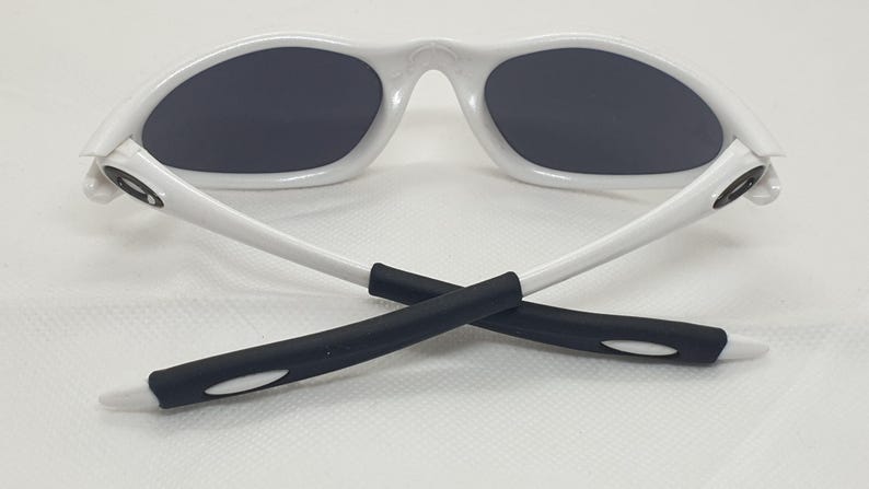 Puede incluir: Gafas de sol blancas con lentes grises oscuras. Las gafas tienen una montura y patillas blancas, con agarres de goma negra en los extremos. Las patillas est&aacute;n cruzadas.