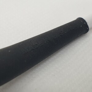 Puede incluir: Primer plano de una secci&oacute;n de goma negra de un cable blanco. La secci&oacute;n negra tiene una superficie ligeramente texturizada y se conecta al cable blanco. El fondo es una superficie blanca lisa.
