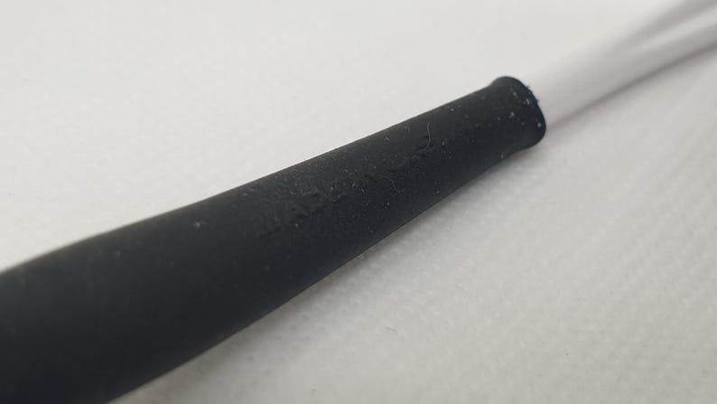 Puede incluir: Primer plano de una secci&oacute;n de goma negra de un cable blanco. La secci&oacute;n negra tiene una superficie ligeramente texturizada y se conecta al cable blanco. El fondo es una superficie blanca lisa.