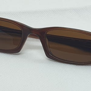 Pode incluir: &Oacute;culos de sol castanhos com arma&ccedil;&atilde;o retangular e lentes coloridas. Os &oacute;culos t&ecirc;m arma&ccedil;&atilde;o e hastes castanhas escuras, e as lentes s&atilde;o de uma cor castanha mais clara. Projetados para prote&ccedil;&atilde;o solar.