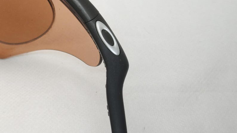 May include: Close-up of black sunglasses with a silver accent on the temple. The lenses are a light brown colour. The sunglasses are set against a white background.