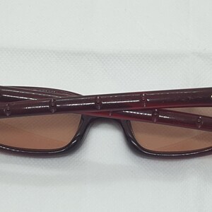 May include: Burgundy sunglasses with a rectangular frame and brown lenses. The temples have a distinctive design with gold accents near the hinges. The sunglasses are folded, revealing the interior and the brand's logo.