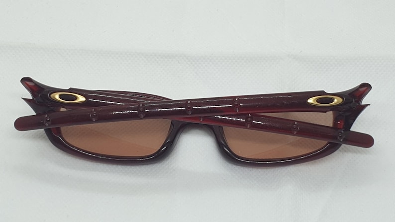 May include: Burgundy sunglasses with a rectangular frame and brown lenses. The temples have a distinctive design with gold accents near the hinges. The sunglasses are folded, revealing the interior and the brand's logo.