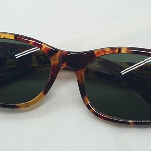 Puede incluir: Gafas de sol con montura de carey y lentes verde oscuro. Las gafas tienen forma rectangular con bordes redondeados. La montura presenta tonos marrones, &aacute;mbar y negros. Un accesorio cl&aacute;sico para proteger los ojos.