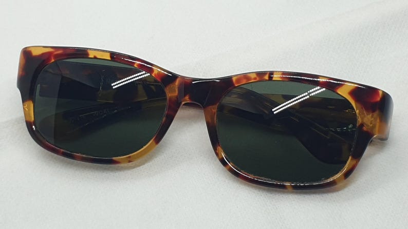 Puede incluir: Gafas de sol con montura de carey y lentes verde oscuro. Las gafas tienen forma rectangular con bordes redondeados. La montura presenta tonos marrones, &aacute;mbar y negros. Un accesorio cl&aacute;sico para proteger los ojos.