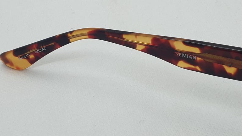 Puede incluir: Primer plano de una patilla de gafas con estampado de carey. La patilla se curva suavemente y presenta el texto "WCAL" y "BOHEMIAN". La paleta de colores incluye tonos marrones, &aacute;mbar y negro.