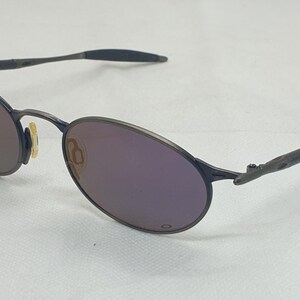 May include: Oval-shaped sunglasses with a dark gray metal frame and purple-tinted lenses. The temples are thin and textured, and the nose pads are a light yellow color. The lenses reflect a gradient of colors.