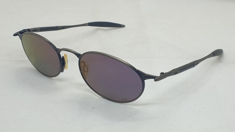 May include: Oval-shaped sunglasses with a dark gray metal frame and purple-tinted lenses. The temples are thin and textured, and the nose pads are a light yellow color. The lenses reflect a gradient of colors.
