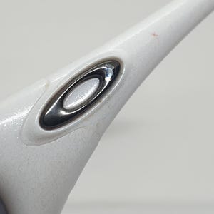 Puede incluir: Primer plano de una patilla de gafas blanca con un logotipo ovalado plateado y negro. La montura tiene un acabado texturizado y moteado. El logotipo es un elemento de dise&ntilde;o prominente.