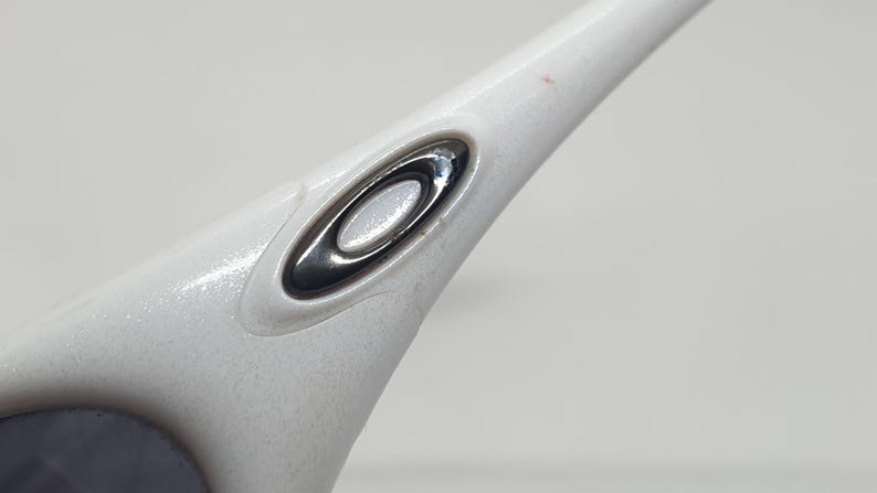 Puede incluir: Primer plano de una patilla de gafas blanca con un logotipo ovalado plateado y negro. La montura tiene un acabado texturizado y moteado. El logotipo es un elemento de dise&ntilde;o prominente.