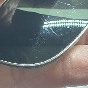 Puede incluir: Primer plano de unas gafas de sol con montura plateada y lentes oscuras. Las lentes tienen ara&ntilde;azos visibles. Las gafas de sol se sostienen en una mano.