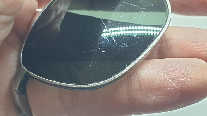 Puede incluir: Primer plano de unas gafas de sol con montura plateada y lentes oscuras. Las lentes tienen ara&ntilde;azos visibles. Las gafas de sol se sostienen en una mano.