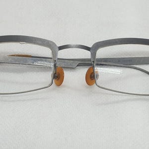 May include: A pair of eyeglasses with a gray metal frame. The lenses are rectangular, and the nose pads are orange. The temples are thin and extend to the sides. The top of the frame is thicker and darker gray.